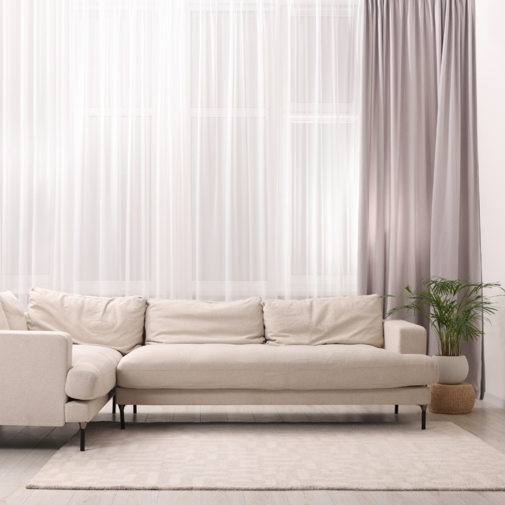 A coastal living room with natural fabric couch, pastel and nature colors. Airy fabrics are used for the curtains giving it a breezy feel. Other natural fiber decorative elements and a potted palm.